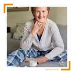 Frau sitzt in gemütlicher Kleidung auf einem Sofa und lächelt in die Kamera