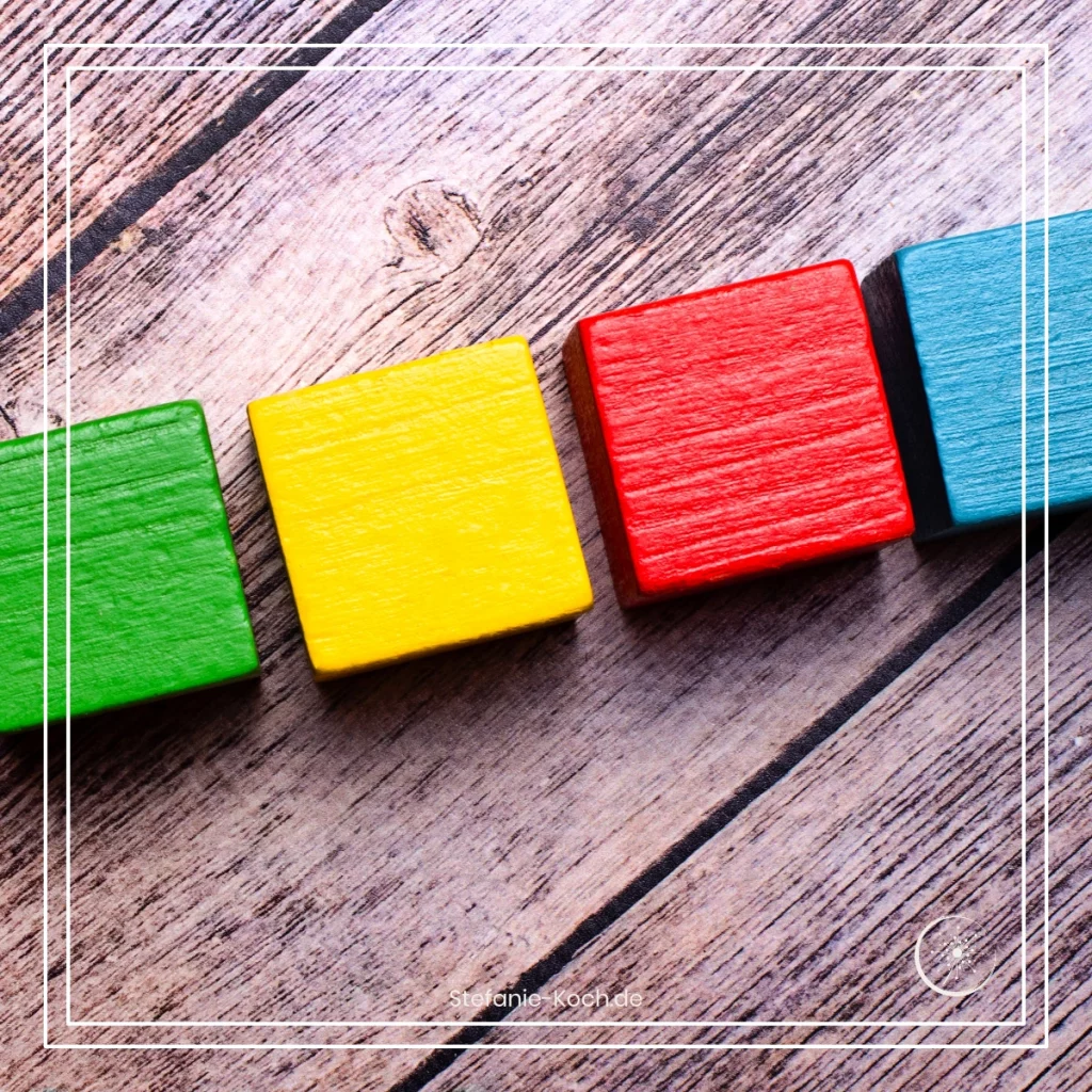 Eine Reihe von vier Bauklötzen auf einer Holzunterlagen - alles ist schwarz-weiß. Ein Bauklotz leuchtet rot, einer leuchter grün, einer gelb und einer blau
