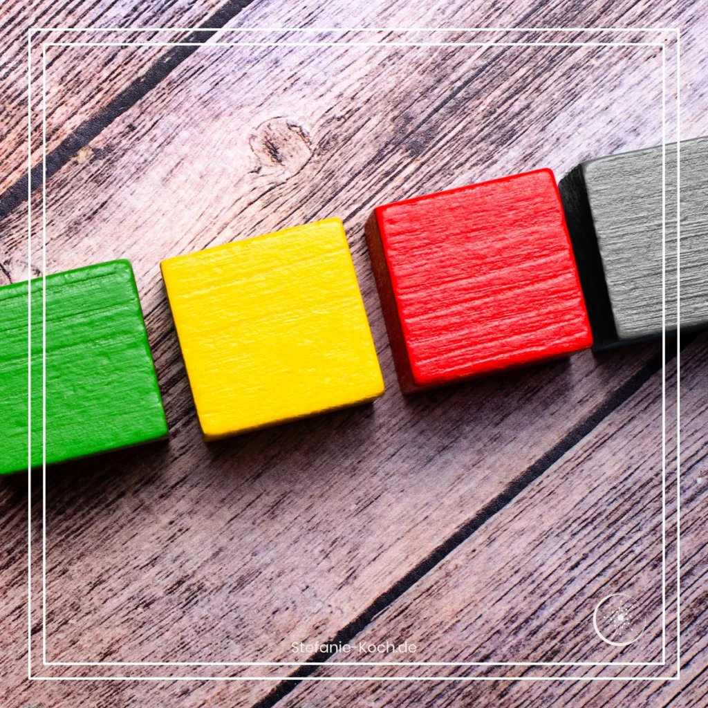 Eine Reihe von vier Bauklötzen auf einer Holzunterlagen - alles ist schwarz-weiß. Ein Bauklotz leuchtet rot, einer grün und einer gelb.