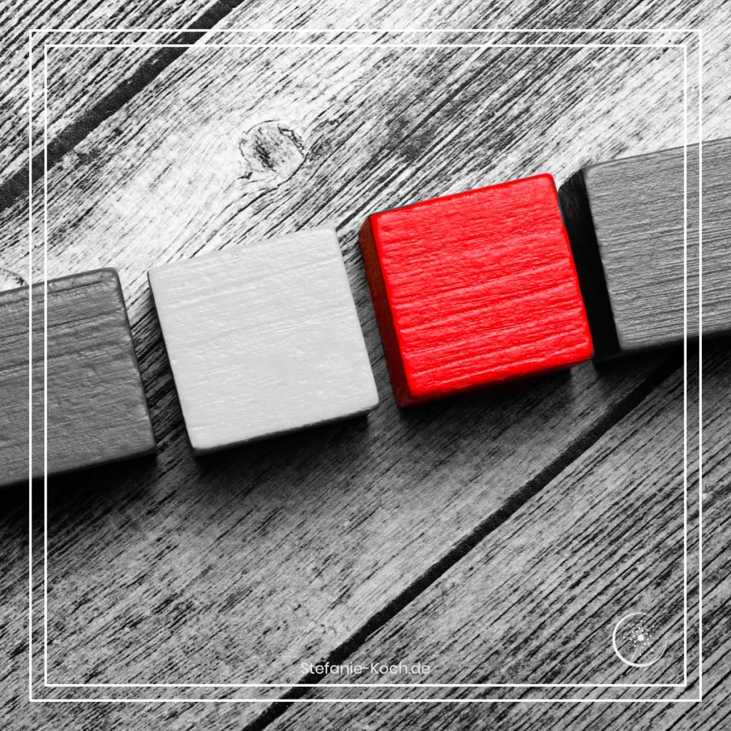 Eine Reihe von vier Bauklötzen auf einer Holzunterlagen - alles ist schwarz-weiß. Ein Bauklotz leuchtet rot.