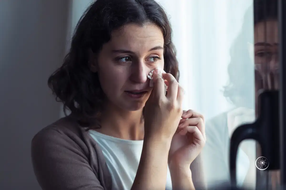 Depression Sinn finden - Eine Frau Anfang 40 steht vor einem FEnster und wischt sich mit einem Taschentuch eine Träne weg