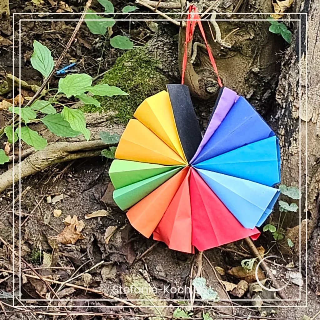 eine Papierblumen in den Farben des Regenbogens liegt auf dem Waldboden