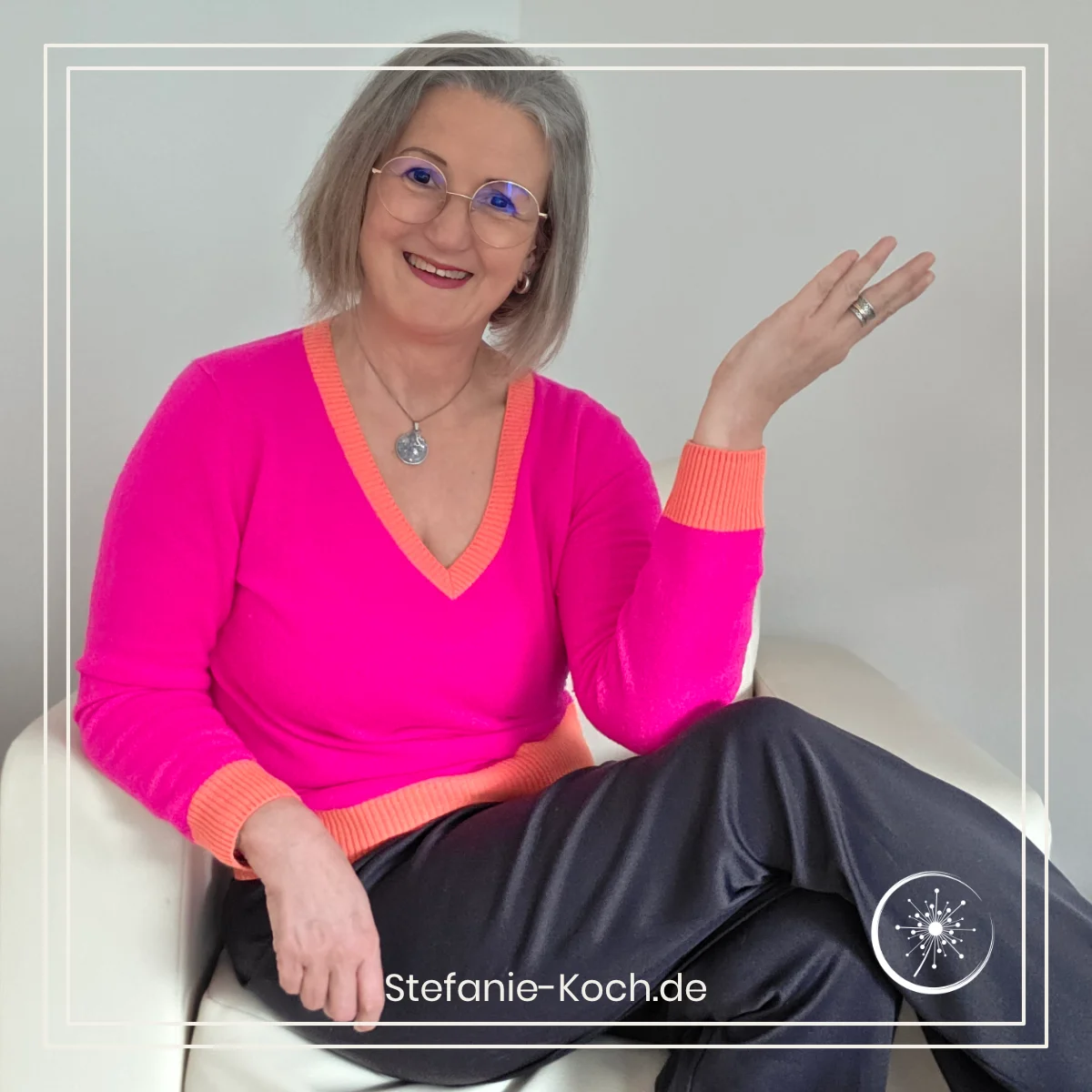 Frau sitzt im Sessel, die linke Hand ist gehoben. Der Blick ist in die Kamera.