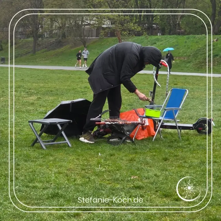 Ein Mann in Regenkelidung auf einer Wiese beim Grillen.