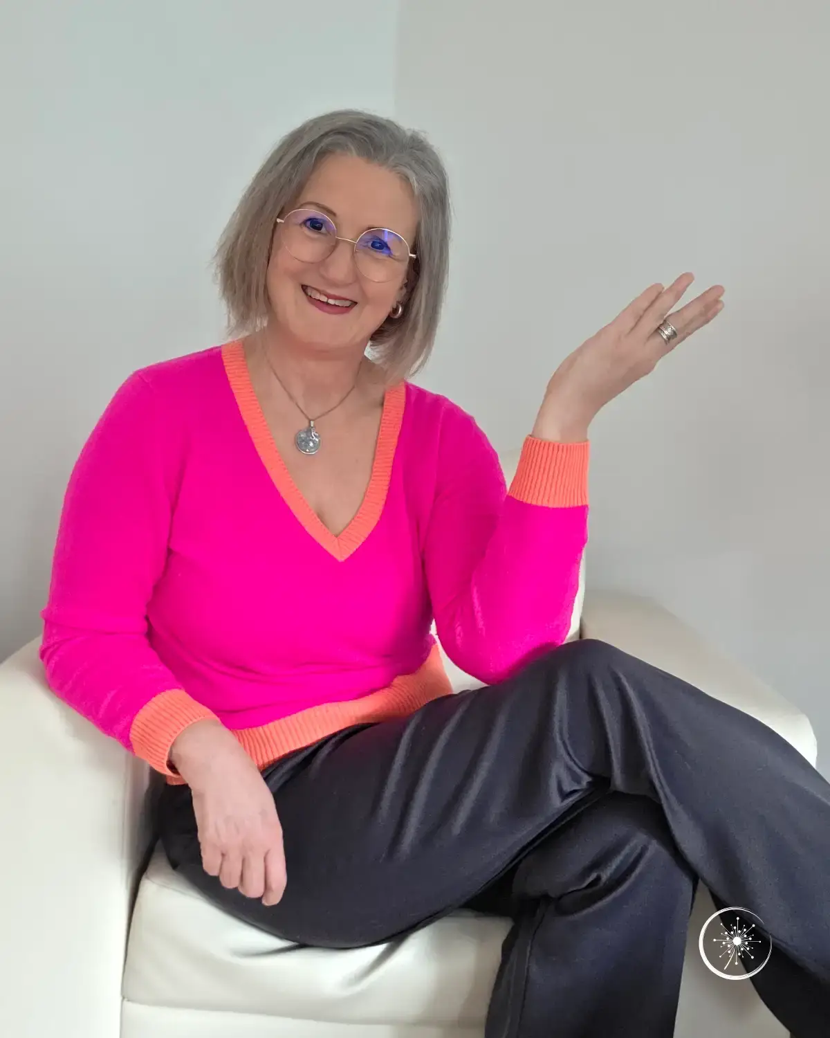 Stefanie Koch sitzt in einem knallig pinkfarbenen Pullover und grauer Stoffhose seitlich auf einem weißen Sessel und lächelt in die Kamera.