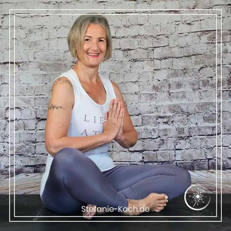 Stefanie Koch sitzt in Yogakleidung auf einer Matte vor einer Steinwand und lächelt in die Kamera.