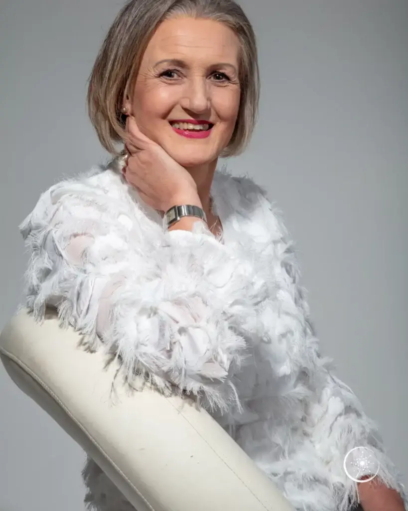 Stefanie Koch sitzt in weißer Fransenbluse und Jeans auf einem weißen Stuhl. Sie lächelt in die Kamera.