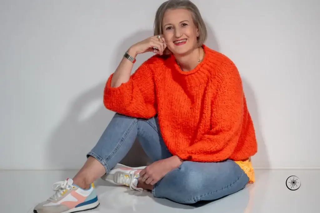 Stefanie Koch sitzt im orangefarbenen Pullover und Jeans auf dem Boden. Sie lächelt in die Kamera.