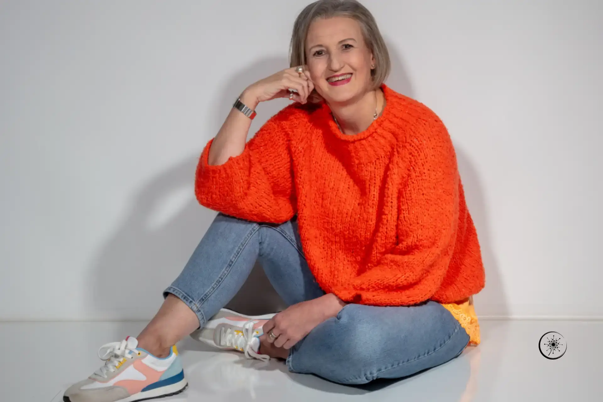 Stefanie Koch sitzt im orangefarbenen Pullover und Jeans auf dem Boden. Sie lächelt in die Kamera.
