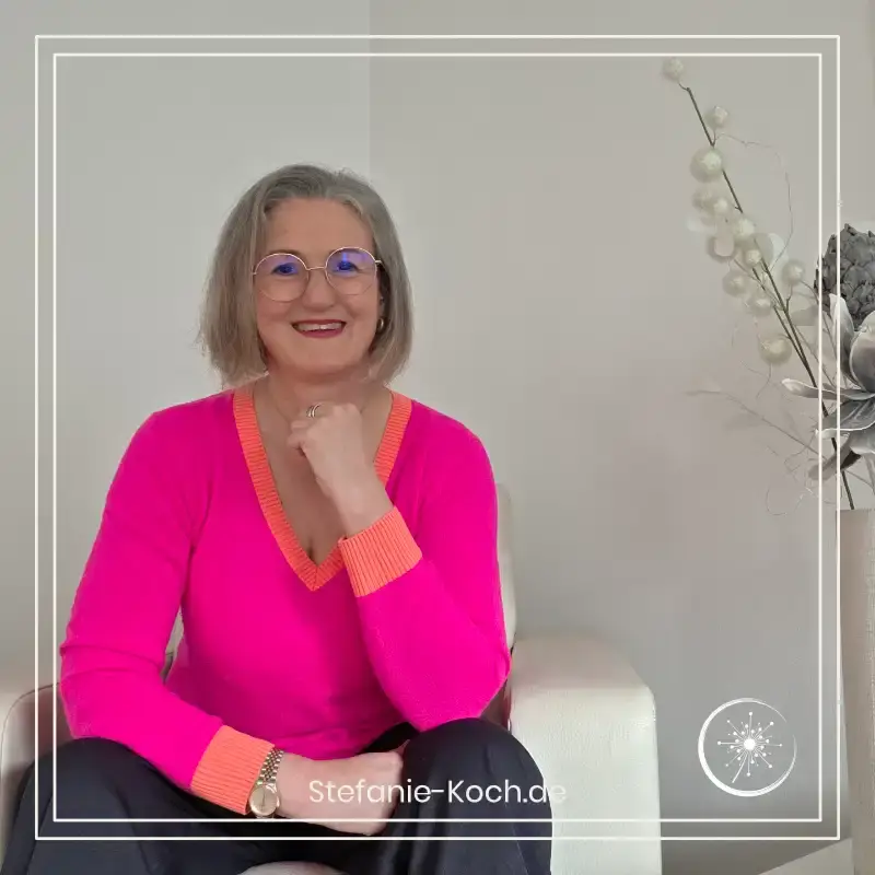 Stefanie Koch sitzt in einem leuchtend pinkfarbenen Pullover frontal auf einem hellen Sessel und lächelt in die Kamera