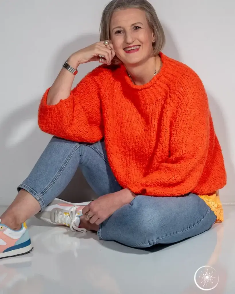 Stefanie Koch sitzt im orangefarbenen Puloover und mit Jeans locker auf dem Fußboden.
