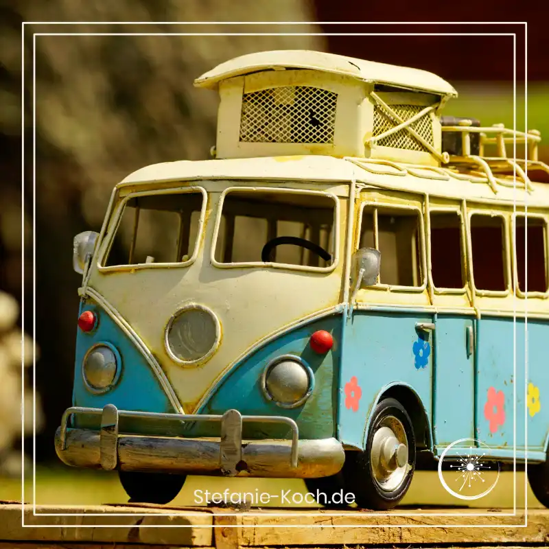 Eine hellblau-weiße Miniatur eines klassischen VW T1 Campingbusses mit bunten Blumen-Aufklebern steht auf einer Holzoberfläche im Freien.
