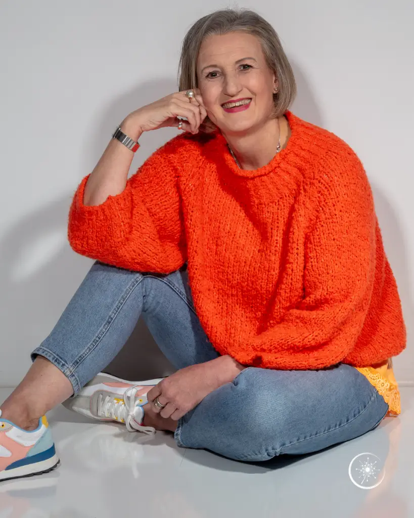 Stefanie Koch sitzt im orangefarbenen Pullover und mit Jeans locker auf dem Fußboden.